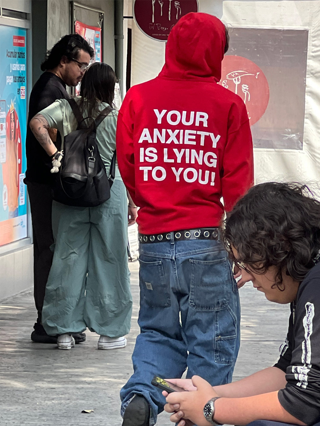 Red hoodie with white text reading "YOUR ANXIETY IS LYING TO YOU" on back, worn with blue jeans and studded belt.