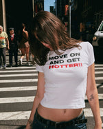 A woman on a city crosswalk wearing a white crop top t-shirt with the text 'MOVE ON AND GET HOTTER!!' printed on the front in black and red lettering.