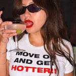 A woman wearing black sunglasses and a white graphic t-shirt with the text 'MOVE ON AND GET HOTTER!!' in black and red lettering. She is holding a coupe glass with a pink cocktail and a lime wedge, set against a dark background.