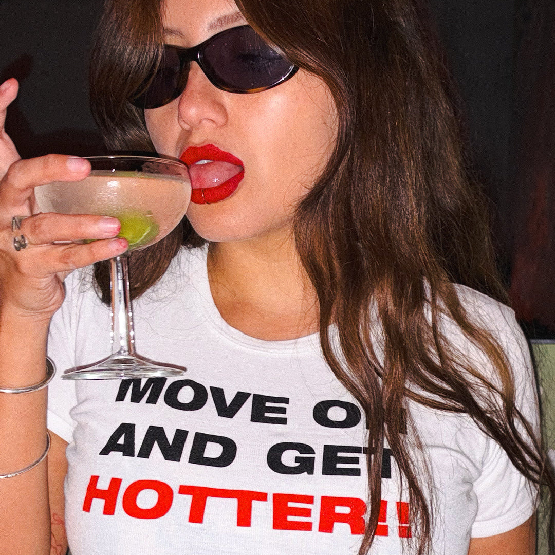 A woman wearing black sunglasses and a white graphic t-shirt with the text 'MOVE ON AND GET HOTTER!!' in black and red lettering. She is holding a coupe glass with a pink cocktail and a lime wedge, set against a dark background.