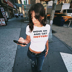 A woman walking on a city sidewalk wearing a white graphic t-shirt with the text 'MOVE ON AND GET HOTTER!!' in black and red lettering, paired with distressed blue jeans.