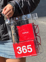 Clear tote bag containing red "Couples 365" pouches and burgundy sunglasses, held by person wearing black jacket.