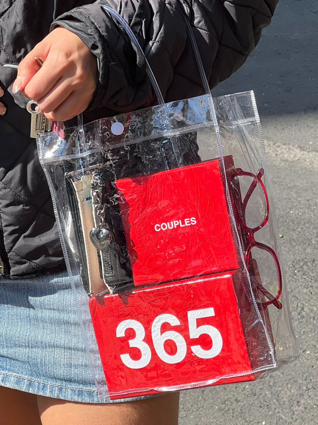 Clear tote bag containing red "Couples 365" pouches and burgundy sunglasses, held by person wearing black jacket.