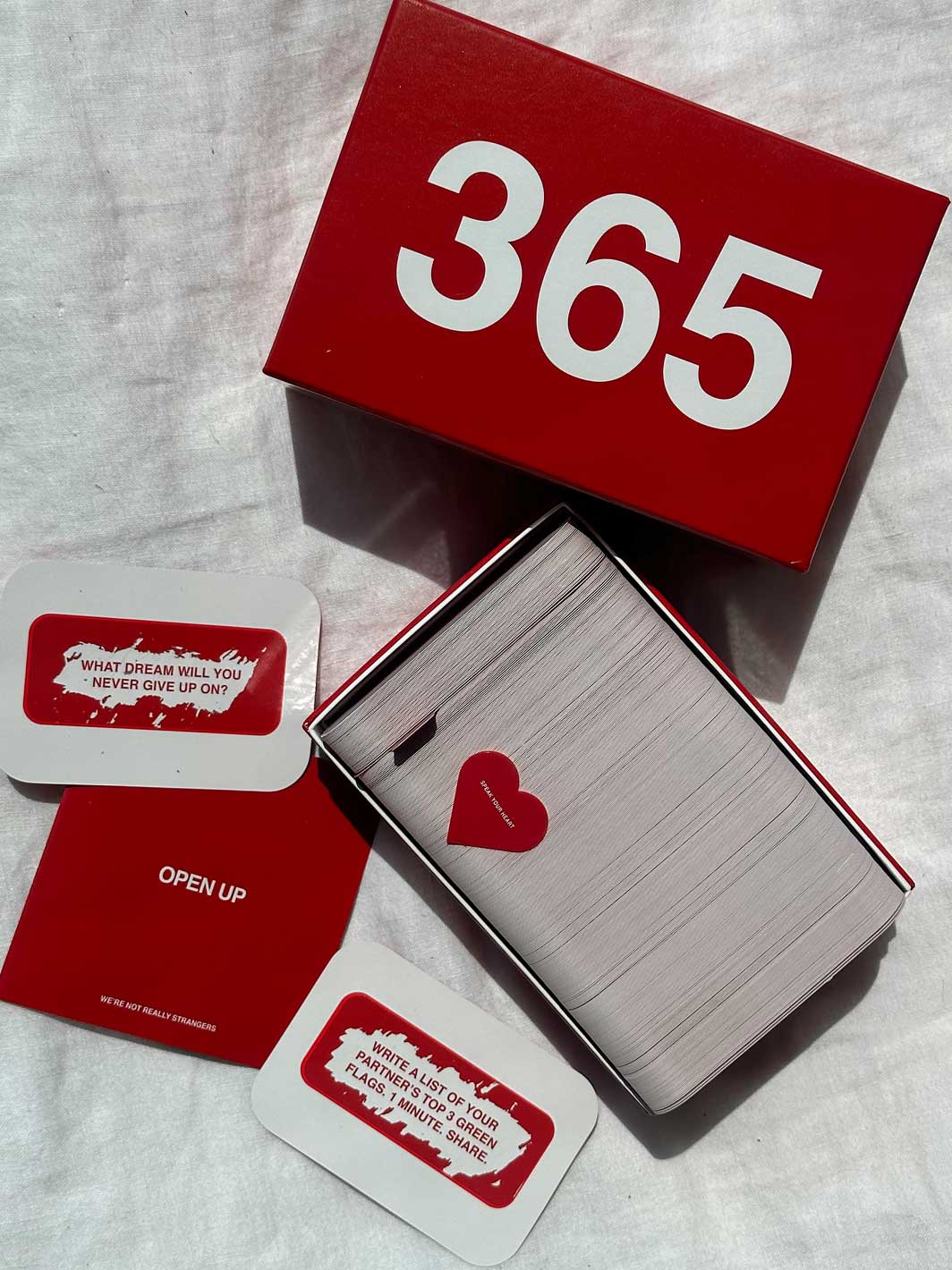 Red box labeled "365" containing white cards with prompts and a red heart charm on a stack of lined pages.