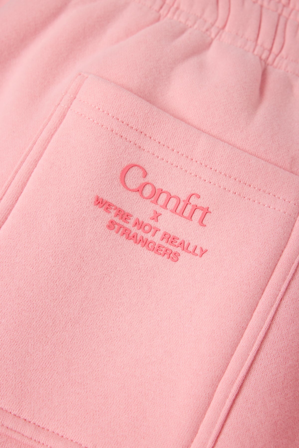 Pink sweatpants with "Comfrt x We're Not Really Strangers" embroidered text on front pocket.