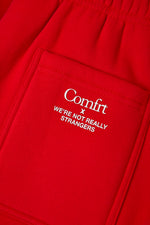 Red sweatpants with white embroidered "Comfrt x We're Not Really Strangers" text and stitched seams.