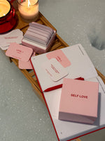 Self Love Game board game with pink and white cards, instruction booklet, and storage box on wooden tray with candles.