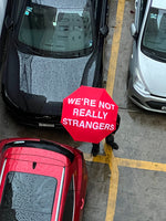 Red umbrella with white text reading "We're Not Really Strangers" held above parked cars.