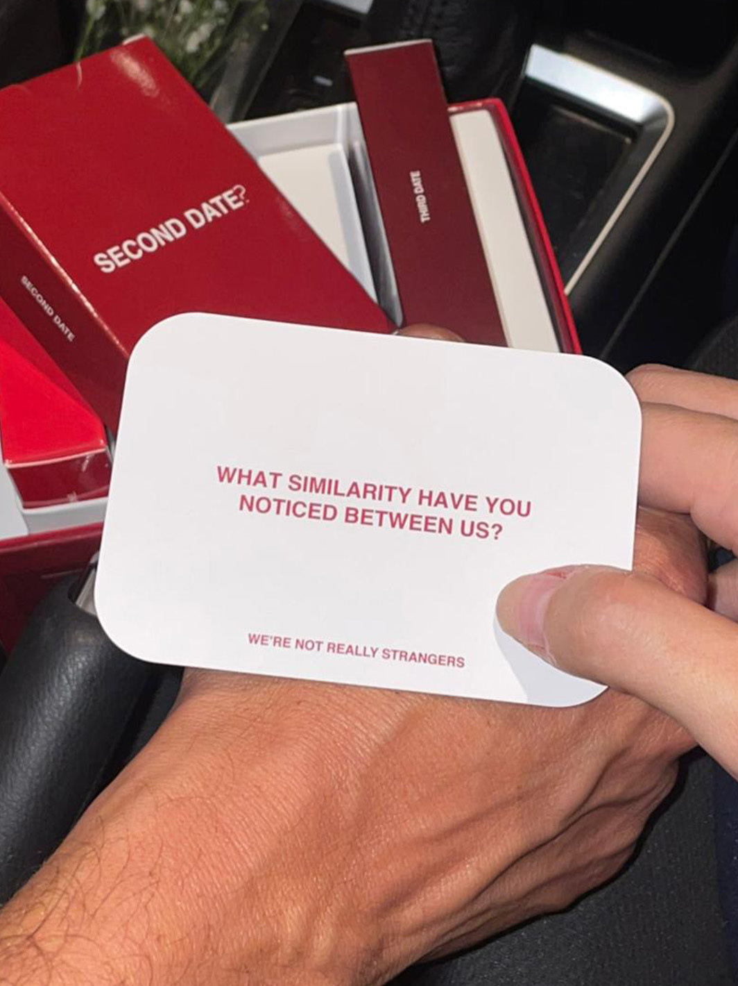 Hand holding white conversation card with pink text next to red "Second Date" boxed kit with white interior.