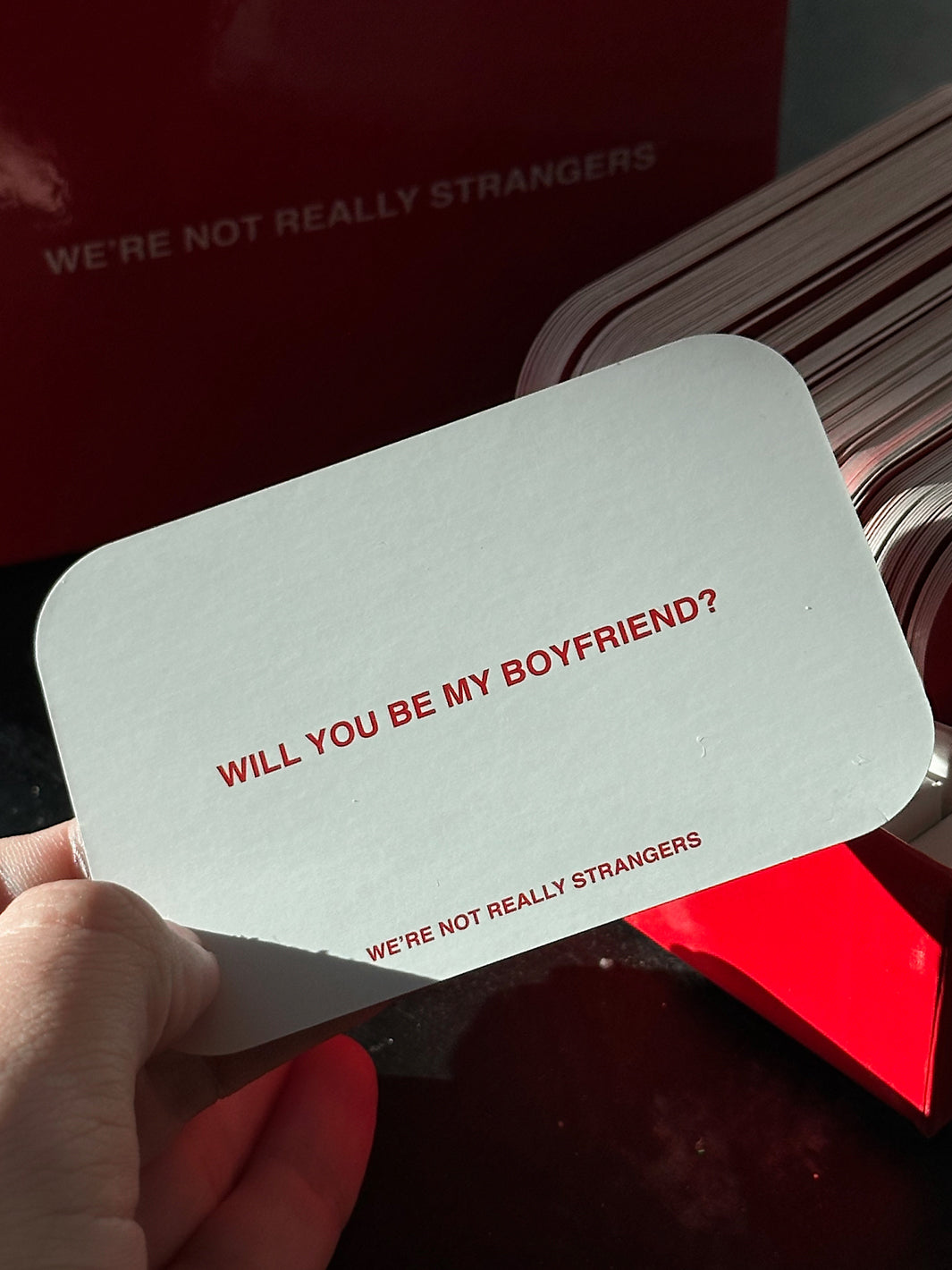 White card with rounded corners displaying "WILL YOU BE MY BOYFRIEND?" in red text, held in hand over red packaging.