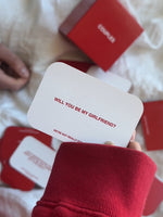White card with red text reading "Will you be my girlfriend? We're not really subtle" held in front of red box packaging.