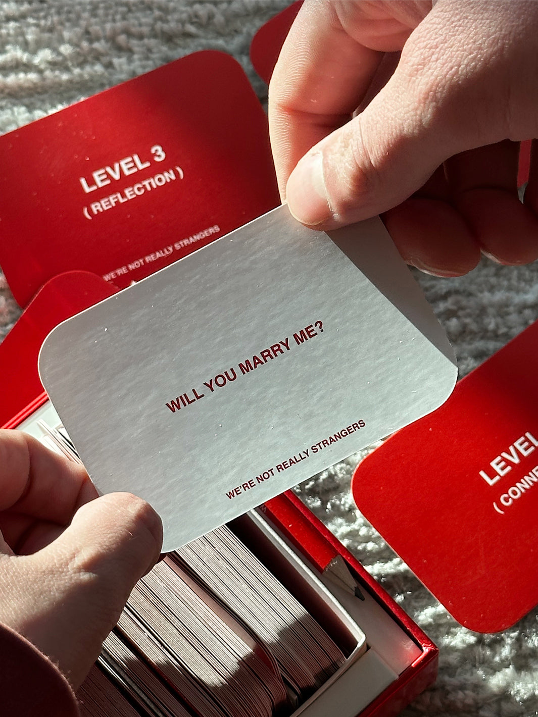 Hand holding white card reading "Will You Marry Me?" with red cards stacked below showing "Level 3 (Reflection)" text.