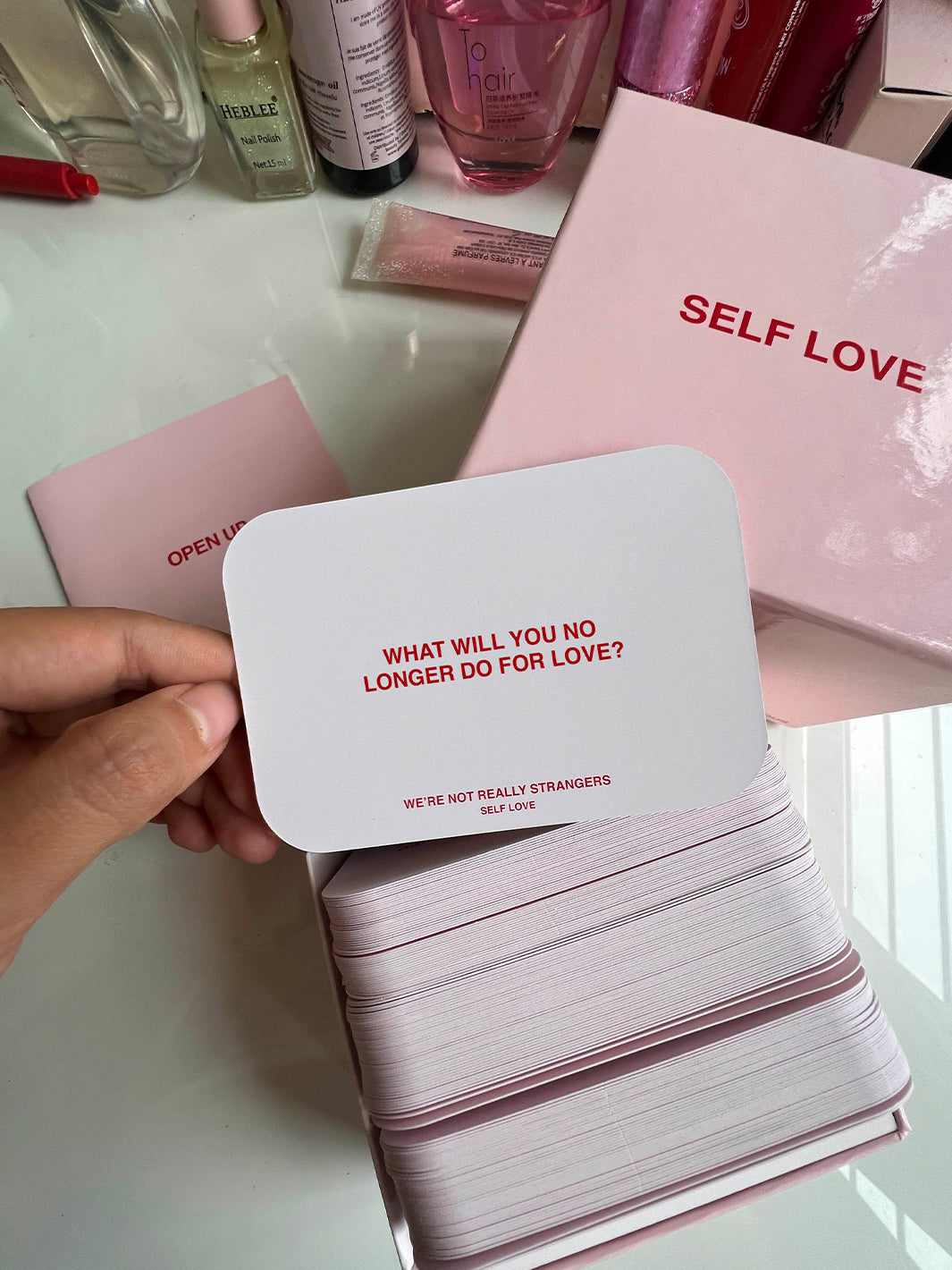 Pink "Self Love" card game with white and red prompt cards displayed in hand, featuring question text and pink box packaging.