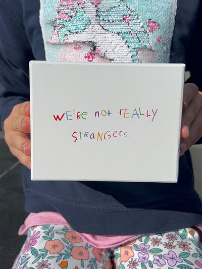 We're Not Really Strangers Kids Edition. Front facing view of kid holding white Kids Edition box reading "We're Not Really Strangers" in rainbow crayon handwriting.