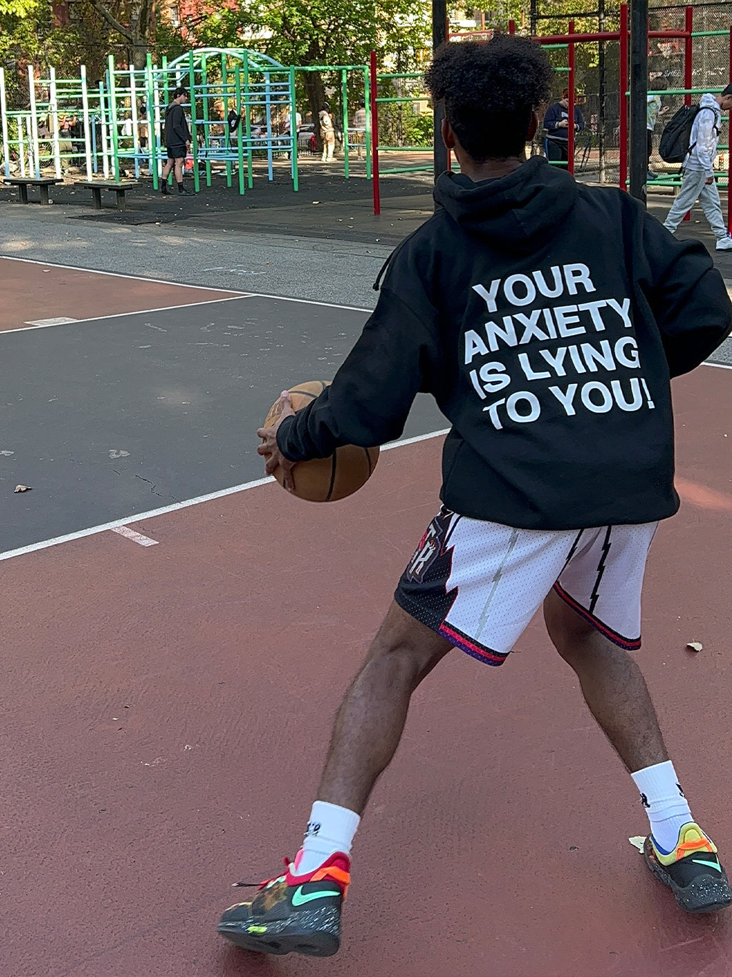 Black hoodie with "YOUR ANXIETY IS LYING TO YOU" text printed on back, worn while playing basketball outdoors.